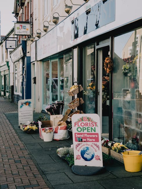 The image depicts the exterior of a florist shop on Belvedere High Street, with a display of fresh flowers and floral arrangements arranged outside the storefront. Potted chrysanthemums and daffodils 