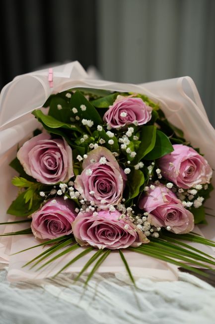 A bouquet of fresh pink roses with delicate, tightly curled petals arranged with green foliage and small white baby's breathe flowers. The bouquet is wrapped in soft white floral paper with a subtle p