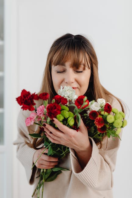 A woman with long, light brown hair and bangs, dressed in a beige knit sweater, holds a colorful floral bouquet close to her face with both hands, gently smelling the flowers. The bouquet features a v