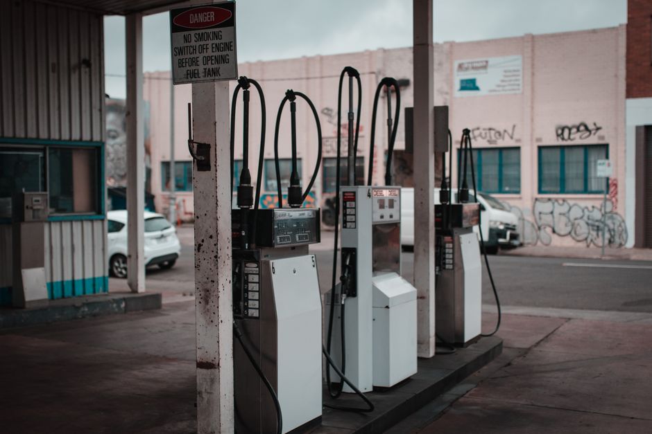 The image depicts a row of four vintage petrol pumps at a gas station, with black hoses dangling from each unit. The pumps are mostly white with some chrome details, situated on a concrete platform un
