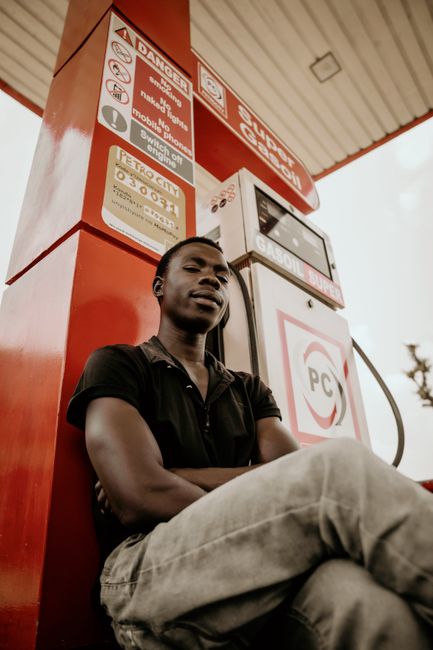 A young man with short, natural hair and a contemplative expression sitting near a red and white petrol pump at a fuel station. He is wearing a black polo shirt and light gray pants. His arms are cros